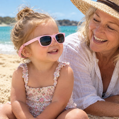 Toddler wearing pink polarised baby sunglasses with strap at the beach beside her grandmother