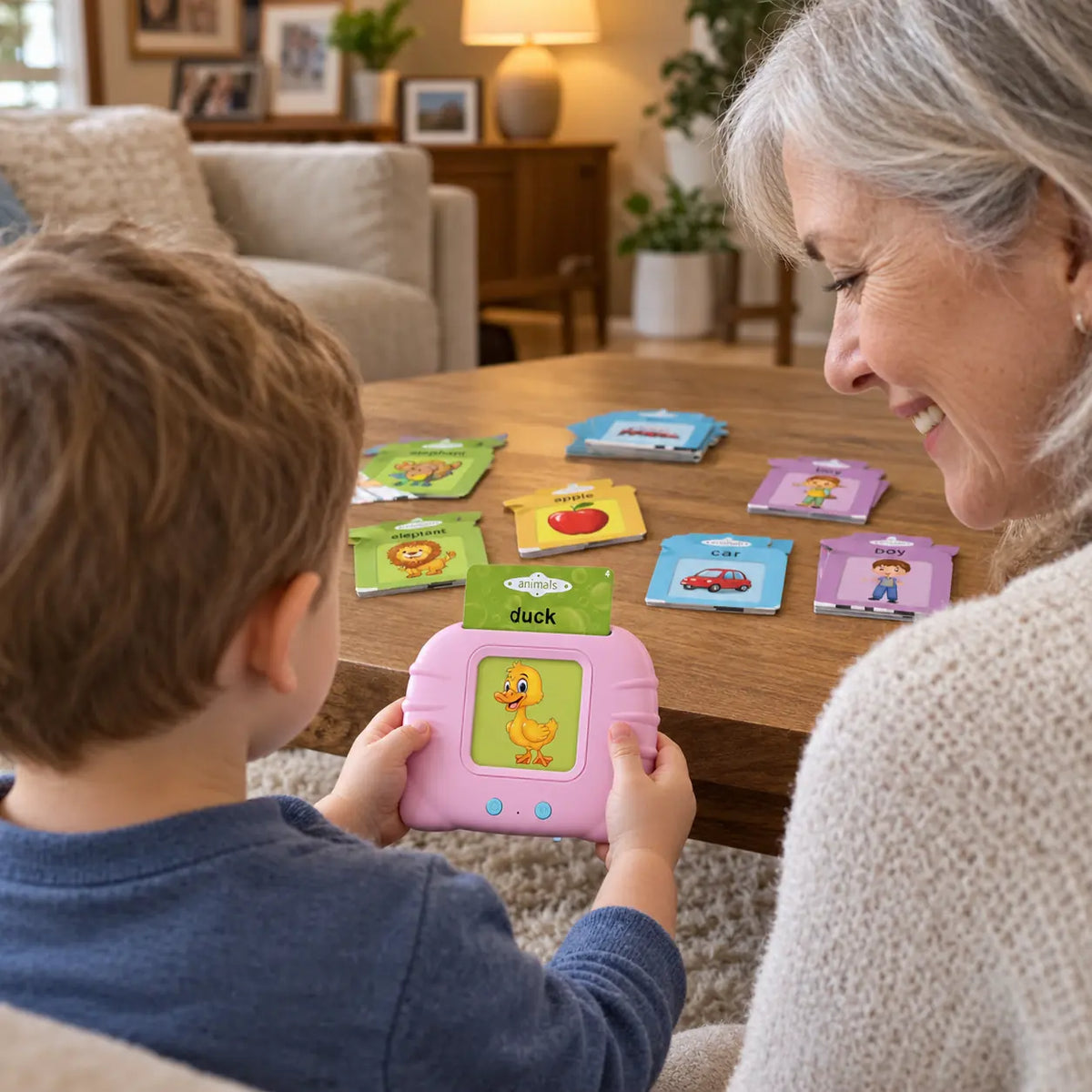 Over-the-shoulder view of a toddler holding the pink audible flash card reader with a duck card, cards spread on the coffee table, grandmother beside him