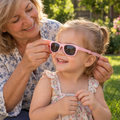 Grandmother putting pink baby sunglasses with strap on her granddaughter in the garden