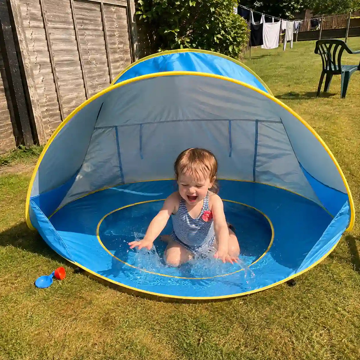 Toddler splashing in the shaded paddling pool of the Little Cubs pop up pool tent on a back garden lawn