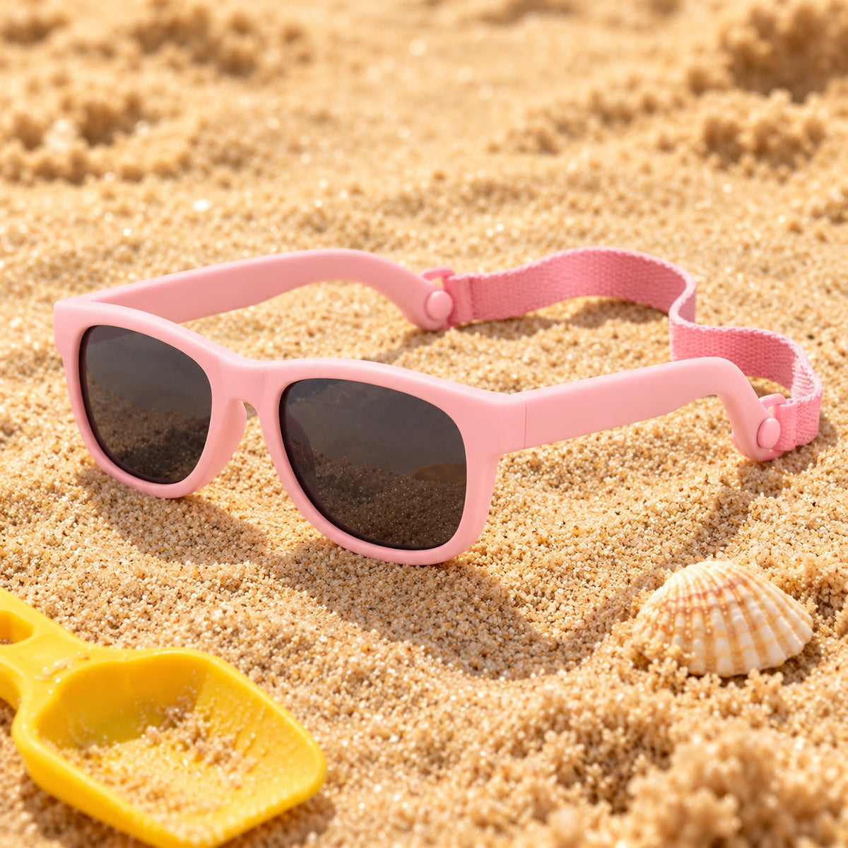 Pink baby sunglasses lying on beach sand beside a shell and toy spade